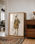 A large framed art print featuring a whimsical illustration of a gazelle dressed as a chef, leaning against a white wall in a modern living room next to a wooden cabinet.