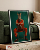 A framed art print leaning against a green sofa in a living room. The print features a zebra wearing a red and green tracksuit sitting on a chair against a dark teal background.