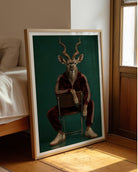 A framed art print leaning against a wall in a sunlit bedroom. The print shows a stylized portrait of a deer-like animal with long curved horns, seated on a chair, wearing a dark red tracksuit and white sneakers against a deep green background.