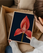 A person holds a framed art print featuring a red orchid flower on a dark navy background, surrounded by brown paper packaging in a cozy indoor setting.