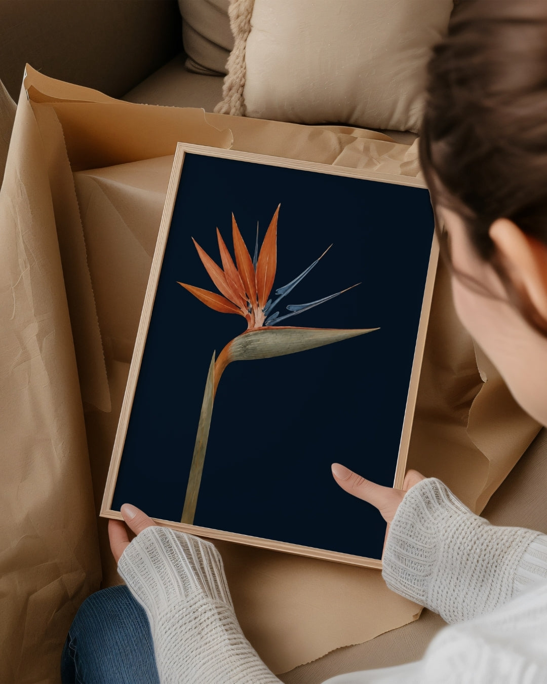 A person holds a framed botanical art print of a bird of paradise flower against a dark navy background. The print is shown inside an open kraft paper box on a sofa.