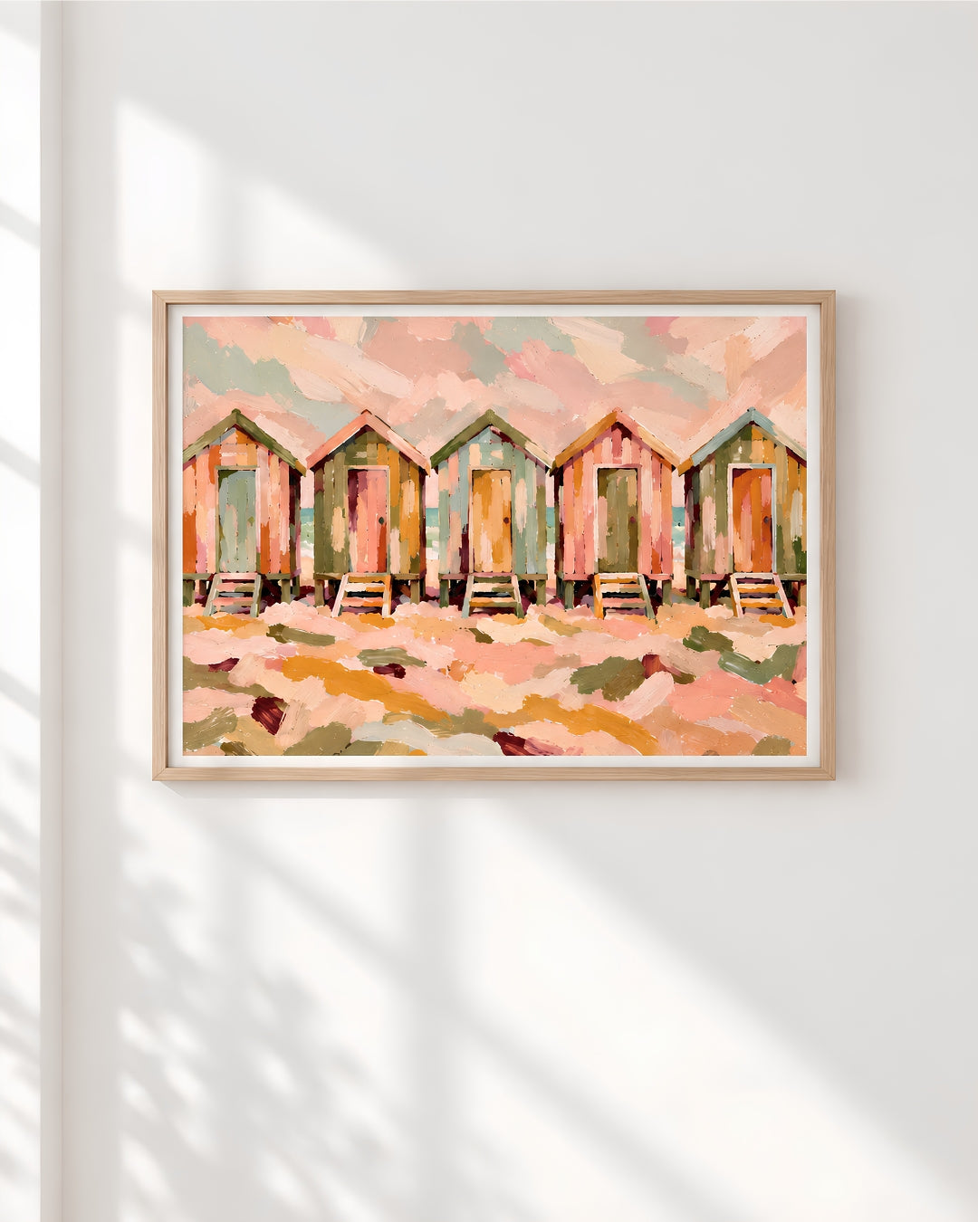 A framed art print depicting a row of beach huts in soft pastel colors, hung on a white wall with decorative shadow patterns cast across it.