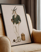 A framed art print featuring a whimsical illustration of a gazelle dressed in vintage menswear playing croquet, leaning against a beige armchair in a cozy room setting.