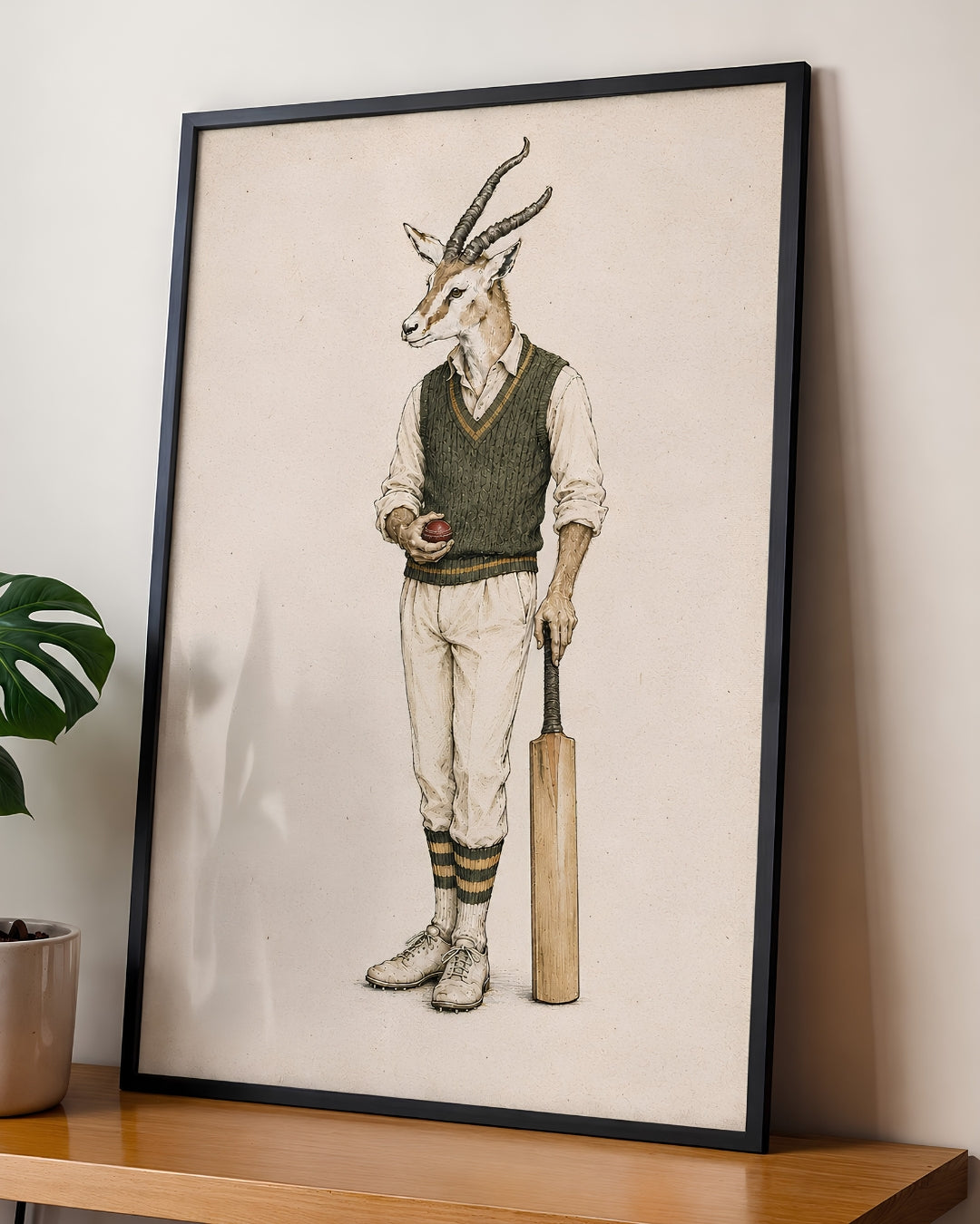 A framed art print featuring a whimsical illustration of an antelope dressed in vintage cricket attire, leaning against a wall on a wooden shelf next to a potted plant.