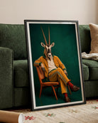 A framed art print leaning against a green sofa in a living room setting. The print features a surreal portrait of a gazelle wearing a yellow tracksuit and sitting on a wooden chair against a dark green background.