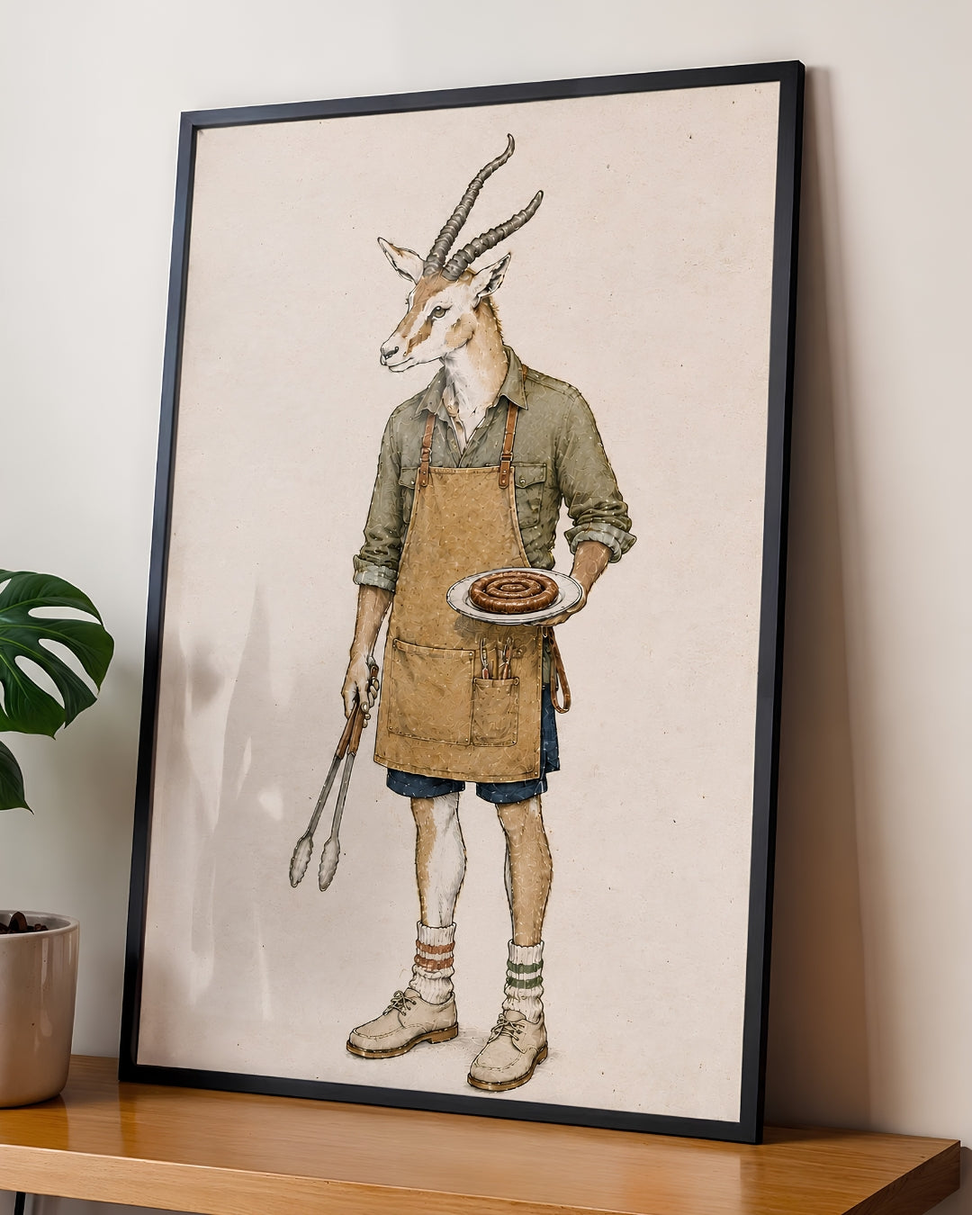 A framed art print featuring an illustration of a gazelle dressed as a chef, leaning against a wall on a wooden shelf next to a potted plant.