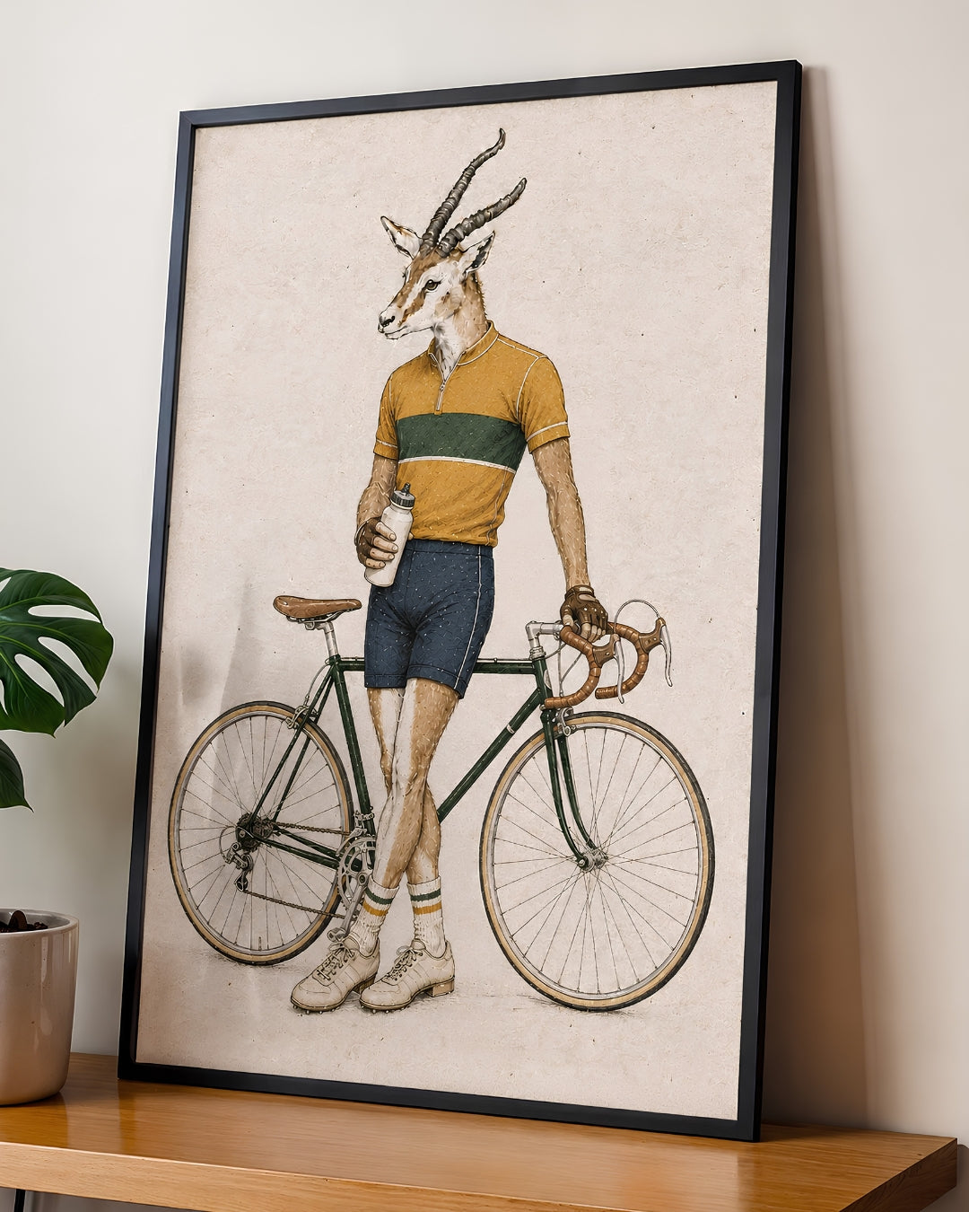 A framed art print featuring a whimsical illustration of a gazelle with antlers riding a vintage road bicycle, wearing a yellow and green striped jersey. The framed print is leaning against a wall on a wooden shelf next to a potted plant.