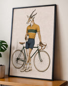A framed art print featuring a whimsical illustration of a gazelle with antlers riding a vintage road bicycle, wearing a yellow and green striped jersey. The framed print is leaning against a wall on a wooden shelf next to a potted plant.