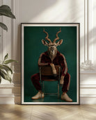 A framed vertical art print featuring a surreal portrait of a kudu-like deer seated on a chair, wearing a dark red tracksuit against a deep teal background. The framed print is displayed leaning against a white paneled wall on a light wood floor, with a green houseplant at the left and soft window shadows across the scene.