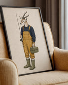 A framed art print featuring a whimsical illustration of a gazelle with antlers dressed in yellow overalls, a navy sweater, and green rain boots while holding a fishing rod and carrying a green box, leaning against a beige armchair in a living room setting.