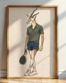 A framed art print featuring a whimsical illustration of an antelope with human characteristics, dressed in a green polo shirt, blue shorts, and holding a tennis racket and tennis ball, displayed on a wooden floor against a light wall.