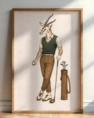 A framed art print featuring a whimsical illustration of a gazelle dressed in golf attire, standing next to a golf bag. The print is housed in a light wood frame and is leaning against a neutral wall on a wooden floor.