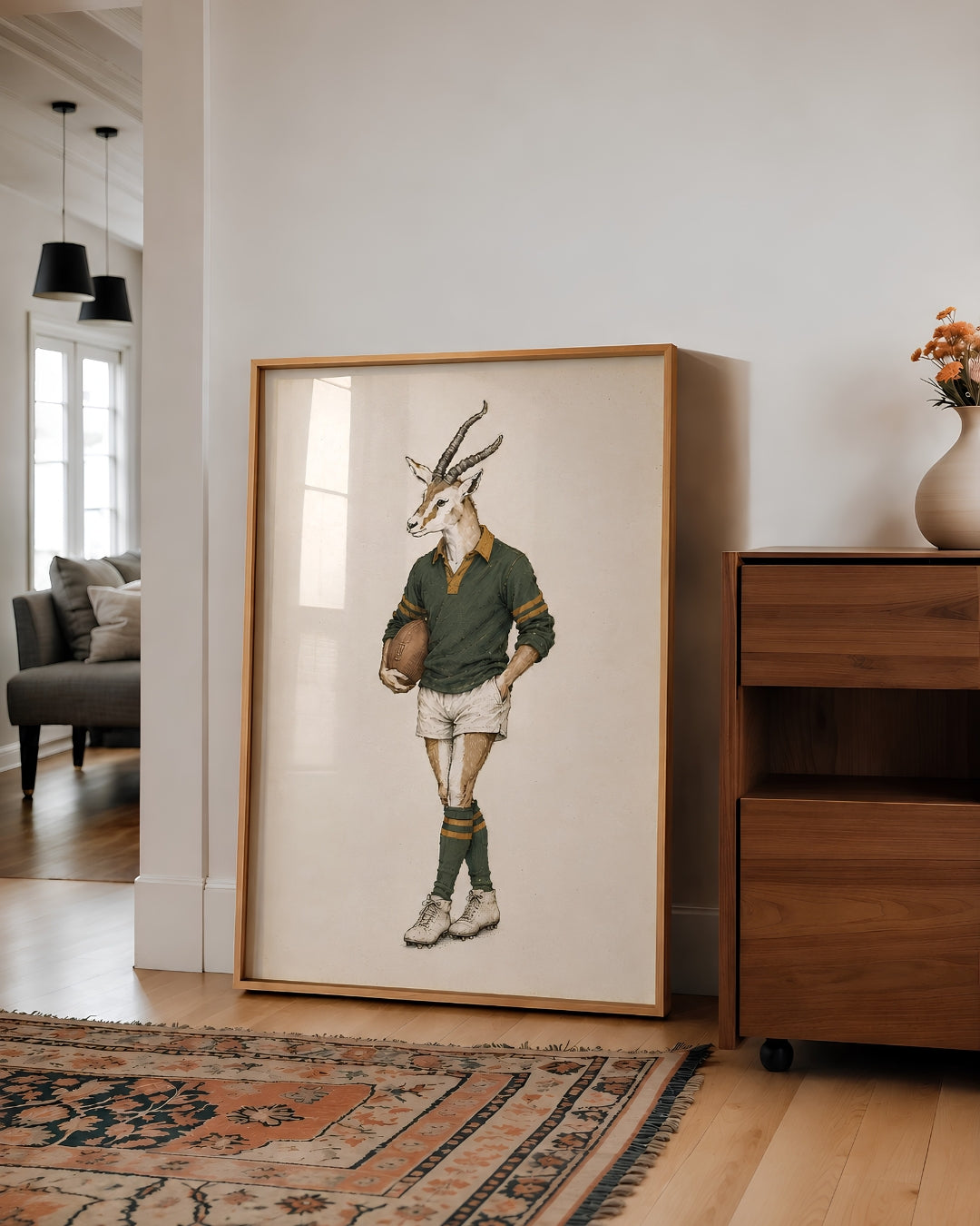 A large framed art print leaning against a white wall in a modern living room. The print features a whimsical illustration of a springbok antelope dressed in a vintage rugby jersey, shorts, socks, and holding a rugby ball.